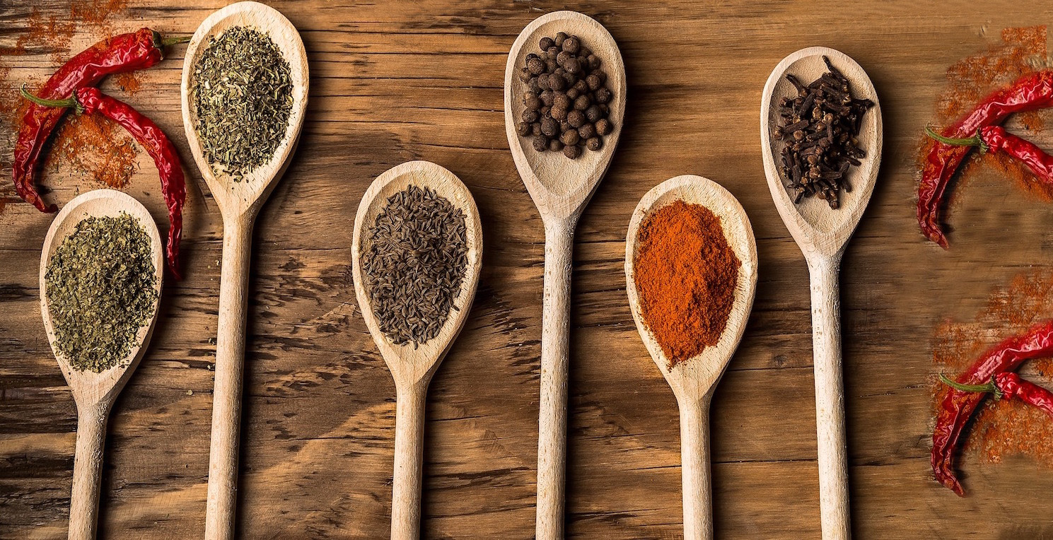 Shows six wooden spoons on a wood plank surrounded by red chili peppers. Each spoon contains a different spice.