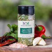 Shows a spice bottle with the My Sage Gourmet label for Aglio Olio e Peperoncino sitting on a wood cutting board and surrounded by an assortment of herbs, salt, chilis, and garlic cloves.