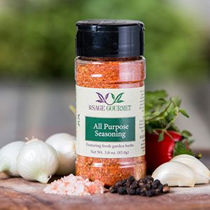 Shows a spice bottle with the My Sage Gourmet label for All Purpose Seasoning sitting on a wood cutting board and surrounded by an assortment of herbs, salt, pepper, onions, and garlic cloves.