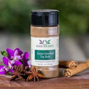 Shows a spice bottle with the My Sage Gourmet label for Asian Smoked Tea Rub sitting on a wood cutting board and surrounded by an star anise, cinnamon, and orchids