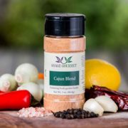 Shows a spice bottle with the My Sage Gourmet label for Cajun Blend sitting on a wood cutting board and surrounded by an assortment of herbs, salt, peppercorns, chilis, garlic cloves, onions, and lemon