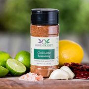 Shows a spice bottle with the My Sage Gourmet label for Chili Lime Seasoning sitting on a wood cutting board and surrounded by an assortment of herbs, salt, chilis, garlic cloves, and citrus fruits