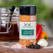 Shows a spice bottle with the My Sage Gourmet label for Chipotle Honey Rub sitting on a wood cutting board and surrounded by an assortment of herbs, salt, pepper, garlic cloves, and citrus fruits