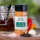Shows a spice bottle with the My Sage Gourmet label for Chipotle Honey Rub sitting on a wood cutting board and surrounded by an assortment of herbs, salt, pepper, garlic cloves, and citrus fruits