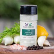 Shows a spice bottle with the My Sage Gourmet label for Dill Seasoning Blend sitting on a wood cutting board and surrounded by an assortment of herbs, salt, pepper, onions, garlic cloves, and lemon