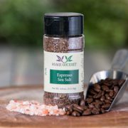 Shows a spice bottle with the My Sage Gourmet label for Espresso Sea Salt sitting on a wood cutting board and surrounded by pink salt and coffee beans