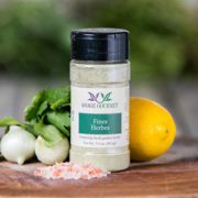 Shows a spice bottle with the My Sage Gourmet label for Fines Herbes sitting on a wood cutting board and surrounded by an assortment of herbs, salt, onion, and lemon