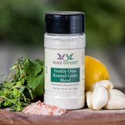 Shows a spice bottle with the My Sage Gourmet label for Freshly Oven Roasted Garlic Blend sitting on a wood cutting board and surrounded by an assortment of herbs, salt, garlic cloves, and lemon
