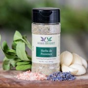 Shows a spice bottle with the My Sage Gourmet label for Herbs de Provence sitting on a wood cutting board and surrounded by an assortment of herbs, salt, garlic cloves, and lavender