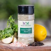 Shows a spice bottle with the My Sage Gourmet label for Lemon Pepper Blend sitting on a wood cutting board and surrounded by an assortment of herbs, salt, peppercorns, garlic cloves, and lemon