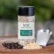 Shows a spice bottle with the My Sage Gourmet label for Summer Garden Grilling sitting on a wood cutting board and surrounded by an assortment of peppercorns, salt, and maple sugar granules