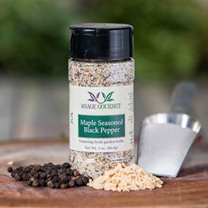 Shows a spice bottle with the My Sage Gourmet label for Summer Garden Grilling sitting on a wood cutting board and surrounded by an assortment of peppercorns, salt, and maple sugar granules