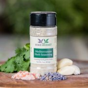Shows a spice bottle with the My Sage Gourmet label for Mediterranean Herb Seasoning sitting on a wood cutting board and surrounded by an assortment of herbs, salt, and various spices