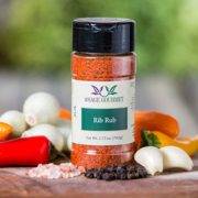 Shows a spice bottle with the My Sage Gourmet label for Rib Rub sitting on a wood cutting board and surrounded by an assortment of herbs, salt, chilis, and various spices