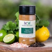 Shows a spice bottle with the My Sage Gourmet label for Sassy Cilantro sitting on a wood cutting board and surrounded by an assortment of herbs, salt, chilis, and various spices