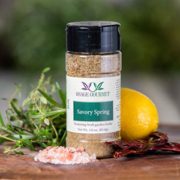 Shows a spice bottle with the My Sage Gourmet label for Savory Spring sitting on a wood cutting board and surrounded by an assortment of herbs, salt, chilis, and various spices