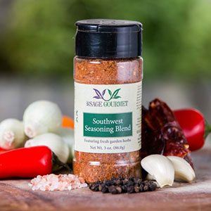 Shows a spice bottle with the My Sage Gourmet label for Southwest Seasoning Blend sitting on a wood cutting board and surrounded by an assortment of herbs, salt, peppercorns, chilis, onions, and garlic cloves.