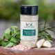Shows a spice bottle with the My Sage Gourmet label for Tuscan Herb Seasoning sitting on a wood cutting board and surrounded by an assortment of herbs, salt, and garlic cloves