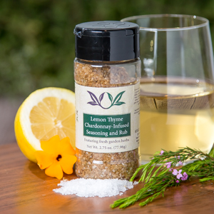 Spice bottle with My Sage Gourmet label for Lemon Thyme Chardonnay Infused Seasoning and Rub set on a cutting board with half an orange, salt, fresh herbs, and a glass of chardonnay wine