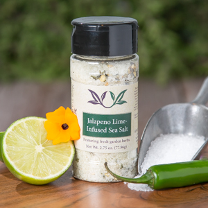 Spice bottle with My Sage Gourmet label for Jalapeño Lime Infused Sea Salt set on a cutting board with half an lime, salt, a jalapeño pepper and a small decorative yellow flower