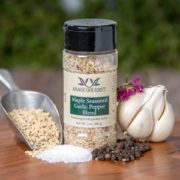 Spice bottle with My Sage Gourmet label for Maple Seasoned Garlic Pepper set on a cutting board with garlic cloves, salt, and peppercorns,