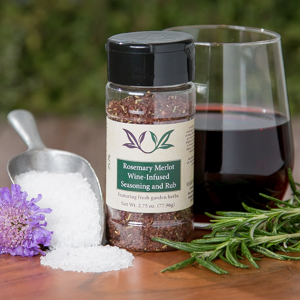 Spice bottle with My Sage Gourmet label for Rosemary Merlot Infused Sea Salt set on a cutting board with salt, fresh herbs, and a glass of merlot wine