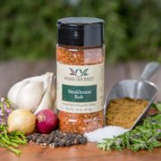 Shows a spice bottle with the My Sage Gourmet label for Steakhouse Rub sitting on a wood cutting board and surrounded by an assortment of herbs, shallots, salt, peppercorns, chilis, and garlic cloves.